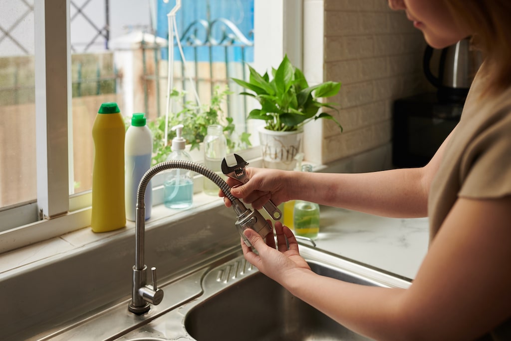Woman checking water faucet with wrench and plants in background.