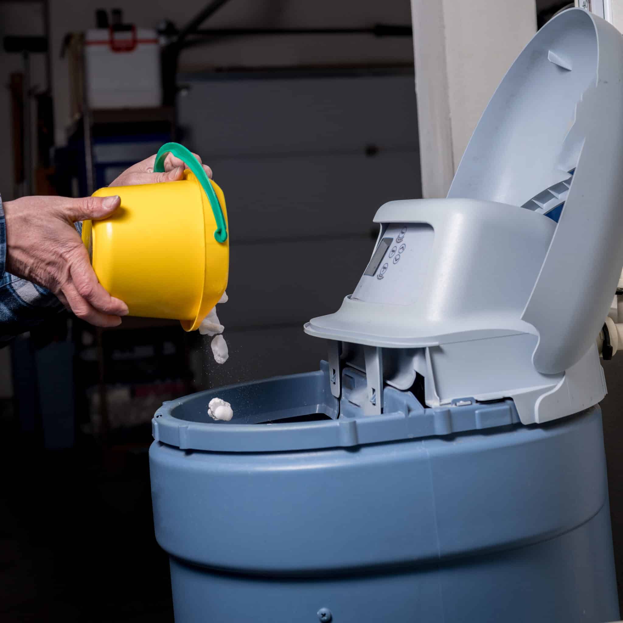 A man pours salt pellets from a yellow bucket into a water softener by Pure Water Solutions.