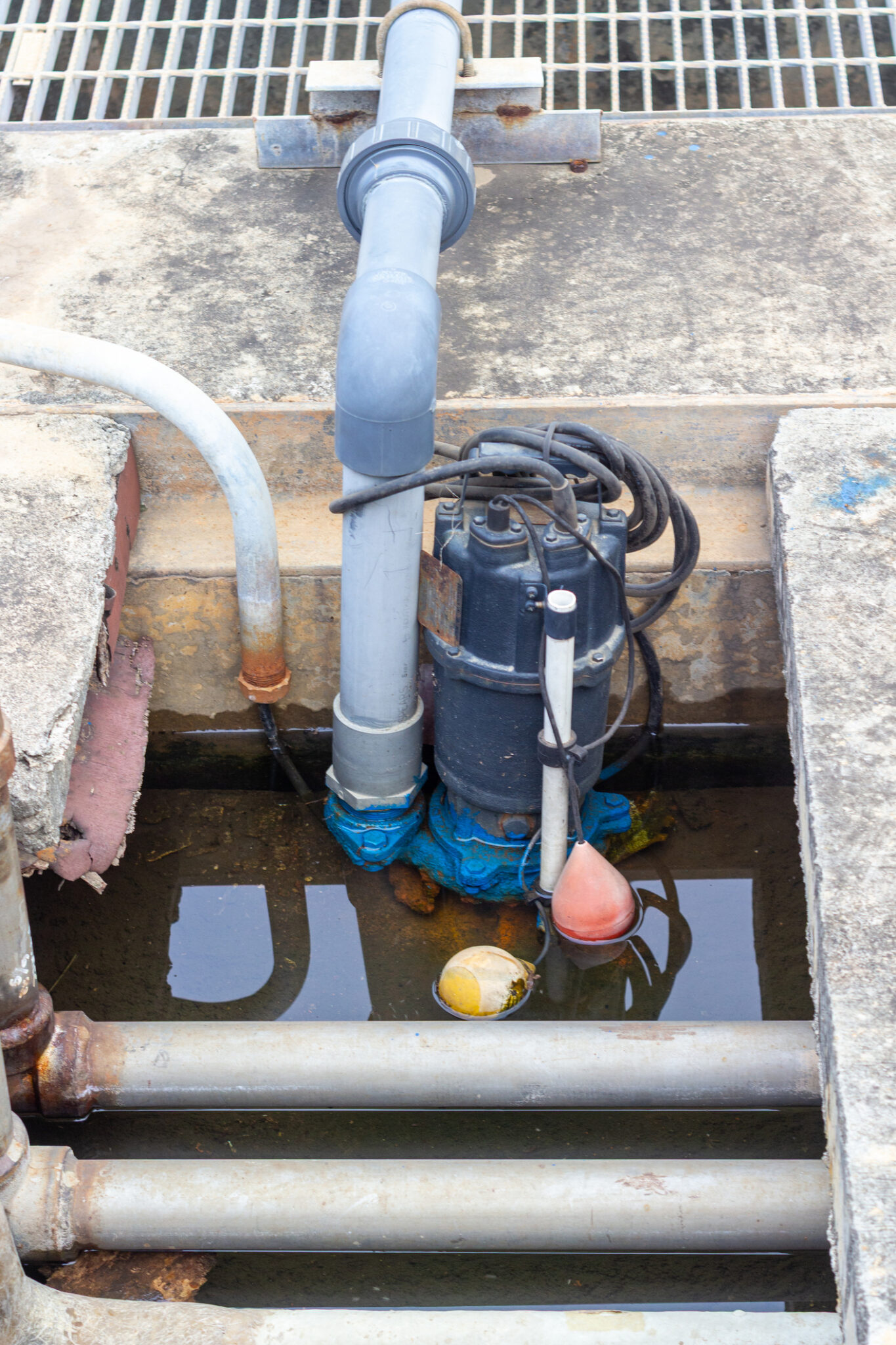 Industrial sewage ejector pump in a pit with exposed pipes, a float switch, and a drainage grate above.